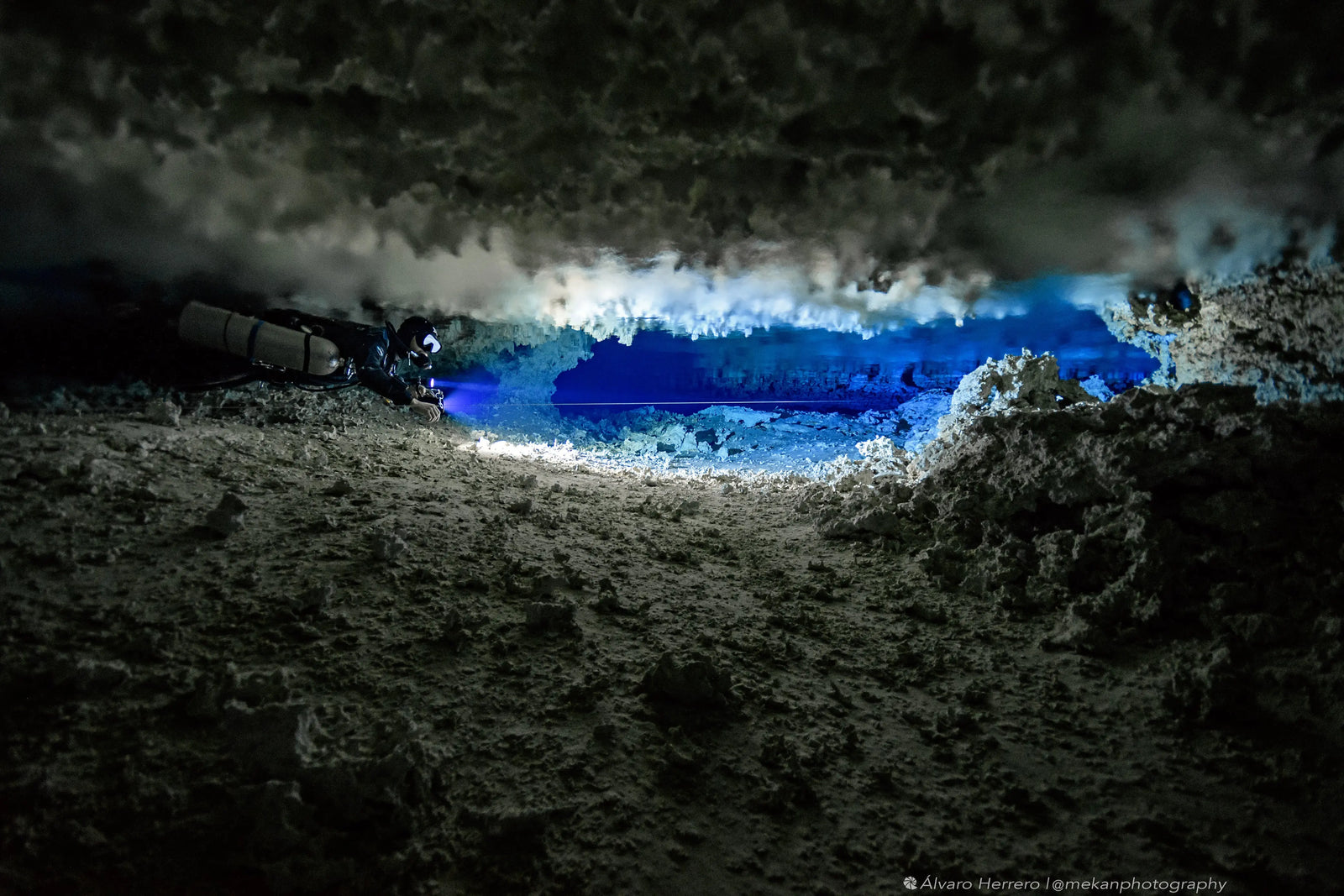 Diving instructor Said el Soueidi navigating a narrow underwater cave passage with a LED SPELEO MKII and a sidemount configuration, illuminating the rough cave walls and blue-lit area ahead.