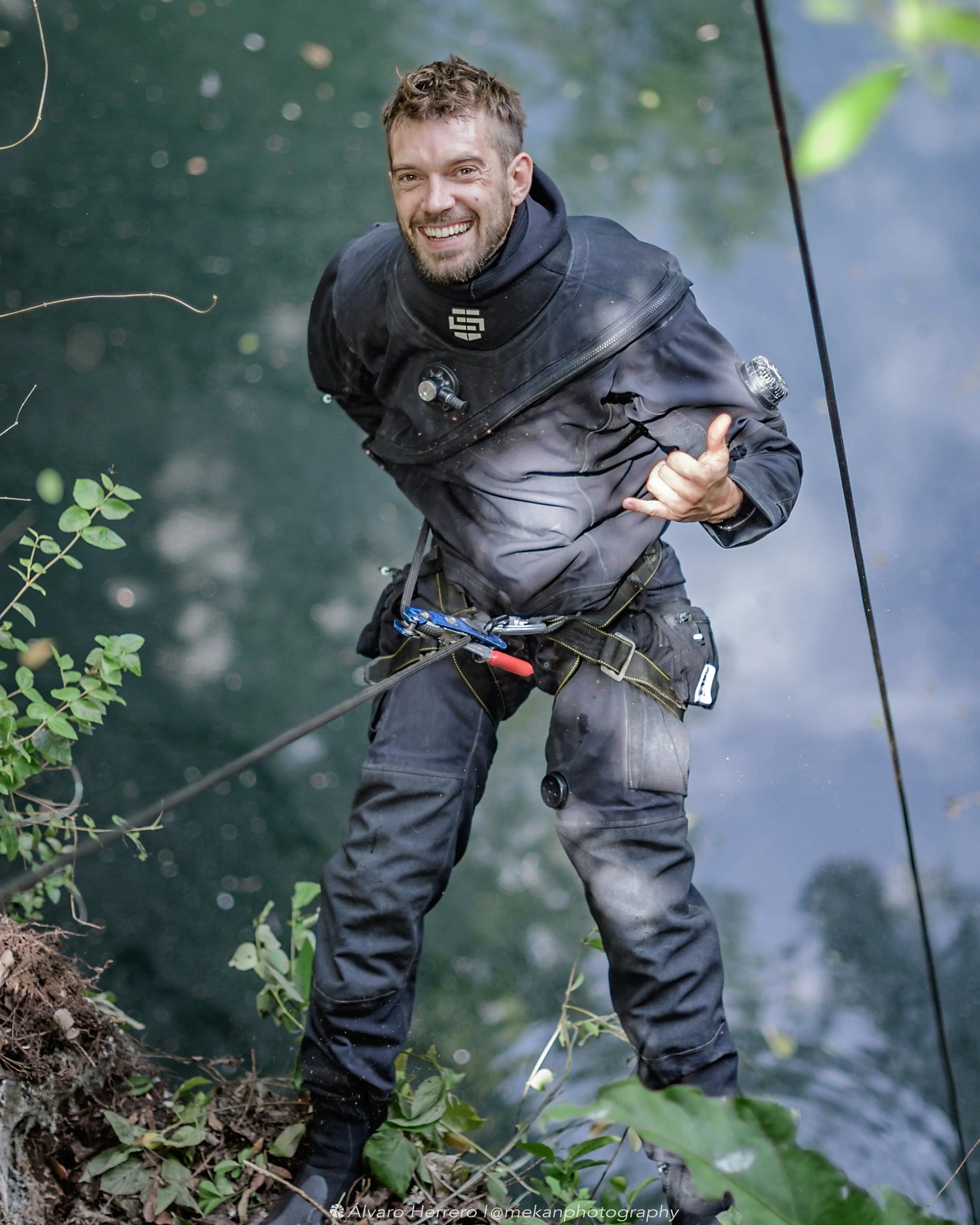 Smiling RAID Certified diving instructor Skanda Coffield-Feith wearing a dry suit, standing by the edge of the water in a tropical setting, with climbing gear and a rope attached.