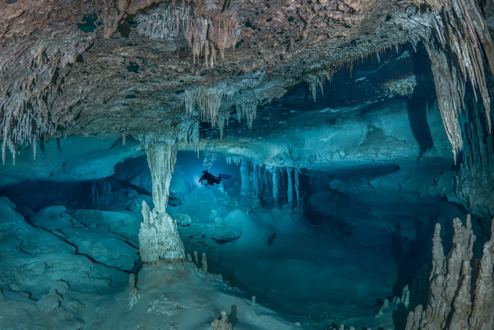 Diving instructor Said el Soueidi using a LED SPELEO MKII to explore a stalactite and stalagmite underwater cave illuminated by blue light, showcasing intricate rock formations and a serene aquatic environment. 